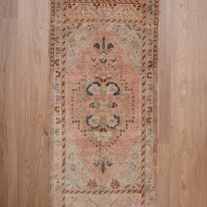 May include: A rectangular, vintage-style rug with a central diamond pattern in soft pink. The rug features intricate floral and geometric designs in shades of beige, brown, and blue. The border is a darker brown with floral motifs. The rug is placed on a light wood floor.