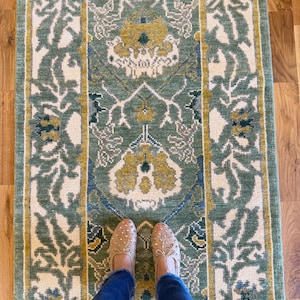 Peut inclure: Un tapis rectangulaire orné d'un motif répétitif de fleurs et de vignes dans des tons de vert sauge, jaune moutarde, crème et bleu marine. Le design comprend un motif central entouré de bordures verticales aux éléments botaniques.