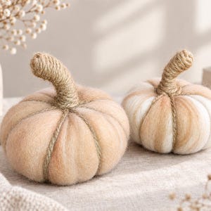May include: Two round decorative pumpkins crafted from soft, felted wool in shades of cream, beige, and light tan. Each pumpkin features a prominent, textured stem wrapped in natural jute twine, with twine segments extending down the sides.