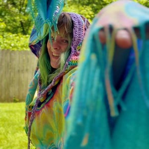 Könnte beinhalten: Ein farbenfroher Batik-Hoodie mit Kapuze und Fransen aus Häkelarbeit. Der Hoodie hat ein lebendiges Wirbelmuster in Gelb-, Blau- und Lilatönen. Die Häkelkapuze ist eine Mischung aus Regenbogenfarben. Die Fransen sind leuchtend türkis.