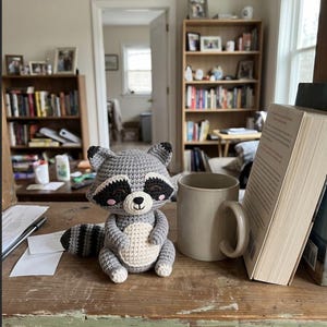 May include: A small, handcrafted crochet raccoon toy featuring gray yarn, a cream-colored belly, black eye patches, and a striped tail. The raccoon is positioned next to a plain beige ceramic mug and an open book on a wooden shelf.