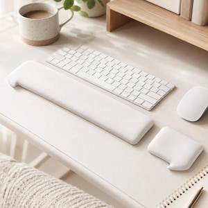 May include: A white ergonomic keyboard wrist rest and a matching white mouse wrist rest. The set is positioned on a desk alongside a white wireless keyboard, a white computer mouse, a speckled ceramic mug, and a spiral-bound notebook.