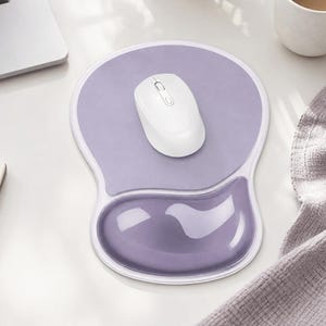 May include: A light purple mouse pad with an integrated gel wrist rest, featuring a smooth surface and a white border. A white wireless computer mouse is positioned in the center of the pad, resting on the flat area above the ergonomic wrist support.