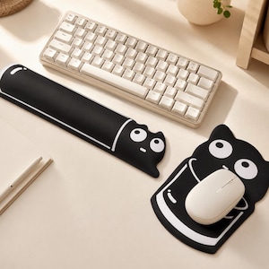 May include: A black and white keyboard wrist rest and a matching mouse pad featuring a cartoon cat design. The wrist rest is long and rectangular, while the mouse pad is shaped like a cat. Both items have white eyes and line details.