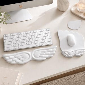 Peut inclure: Un repose-poignet pour clavier et un tapis de souris assorti, tous deux blancs et en forme d'ailes d'ange stylisées. Le repose-poignet est allongé et incurvé, tandis que le tapis de souris présente un bord supérieur arrondi avec des détails d'ailes.