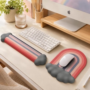 Peut inclure: Un repose-poignet pour clavier et un tapis de souris arborant un motif arc-en-ciel avec des rayures rouges, orange, roses et bleues. Les deux articles ont des extrémités en forme de nuages gris foncé. Le tapis est arqué et le repose-poignet est long.