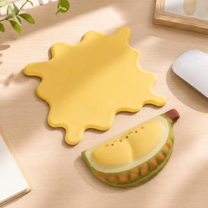 Peut inclure: Un tapis de bureau jaune en forme de tache irrégulière accompagné d'un repose-poignet en forme de croissant arborant un motif de tranche de fruit souriante avec une peau verte, un intérieur jaune clair et des petites marques blush orange.