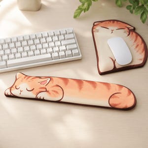 May include: A long, rectangular keyboard wrist rest and a matching mouse pad, both featuring a printed illustration of a sleeping orange tabby cat with closed eyes and dark stripes. The mouse pad is shaped like a sitting cat, and a white mouse sits on it.