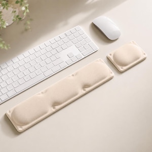 May include: A beige ergonomic keyboard wrist rest and a matching rectangular mouse wrist rest, positioned alongside a white wireless keyboard and a white computer mouse. The wrist rests feature a soft, textured surface with contoured padding.