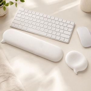 Puede incluir: Un teclado de ordenador blanco, un ratón inalámbrico blanco, un reposamuñecas rectangular largo de color blanco y un pequeño soporte redondo blanco para la muñeca del ratón. Los reposamuñecas tienen una superficie lisa con una pequeña pestaña puntiaguda.