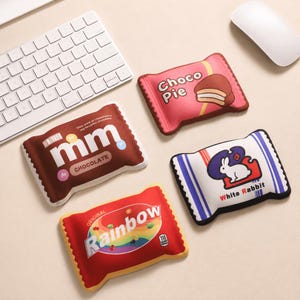 Peut inclure: Quatre coussins rectangulaires souples en forme d'emballages de bonbons, arborant des graphismes colorés de Choco Pie, mm chocolate, Rainbow et White Rabbit. Chaque coussin affiche son nom de marque et ses images, avec des bords festonnés.