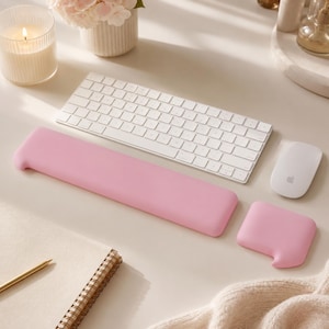 Op de afbeelding: Een lichtroze rechthoekige polssteun voor een toetsenbord en een bijpassende vierkante polssteun voor een muis met afgeronde hoeken. Deze effen siliconen accessoires liggen voor een wit draadloos toetsenbord en een witte computermuis.
