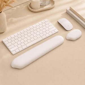 May include: A white wireless keyboard, a white computer mouse, a white ergonomic wrist rest, and a small white pebble-shaped object arranged on a beige surface. A ribbed vase with dried grass and a small tray with a candle are also visible.