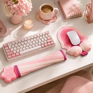 Op de afbeelding: Een mechanisch toetsenbord met witte en roze toetsen, een lange roze polssteun met een stervormig uiteinde en een roze muismat met een witte draadloze muis. De set bevat een roze mok, een roze notitieboek en een vaas met roze bloemen.