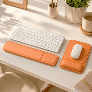 May include: A white wireless keyboard, a white computer mouse, a long rectangular orange wrist rest, and a rectangular orange mouse pad with an integrated wrist support. The items are arranged on a desk surface with a plant and a mug nearby.