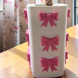 May include: A white square planter decorated with multiple raised pink bows on its sides. A plant with dark purple and green leaves and a speckled stem grows out of the top of the planter. The planter has a smooth finish and a clean, modern shape.