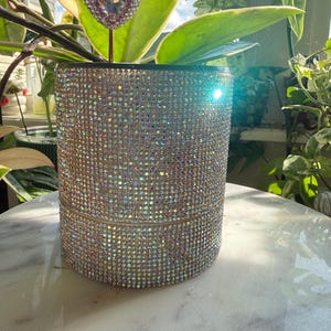 May include: A cylindrical plant pot covered in iridescent, light-reflecting rhinestones. The exterior surface is entirely encrusted with small, shimmering crystals that create a multi-colored sparkle. A small heart-shaped rhinestone pick is visible.