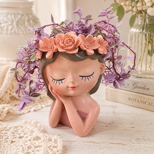 May include: A resin planter shaped like a sleeping face with closed eyes and long lashes. The figure wears a crown of peach-colored sculpted flowers. A trailing purple and green plant grows from the top of the head, and the figure rests its chin on one hand.