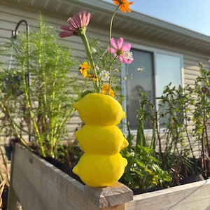 May include: A bright yellow vase shaped like a stack of three lemons, containing a small bouquet of colorful flowers including pink, orange, and white blossoms with green stems, positioned outdoors in natural sunlight.