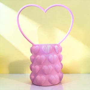 Peut inclure: Un organisateur de bureau cylindrique rose clair présentant un motif répétitif en forme de cœur avec des textures striées verticales. Une grande anse fine en forme de cœur s'étend vers le haut depuis le bord supérieur du récipient.