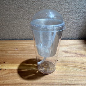 May include: A clear plastic tumbler with a dome lid and a removable silver glitter-infused inner liner. The cup features a tapered cylindrical shape with a smooth exterior surface and a matching clear straw extending from the base of the inner insert.