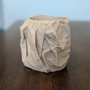 May include: A cylindrical vessel with a light beige, matte finish, featuring a textured surface that mimics the appearance of crumpled paper. The object has an open top and a structured, irregular exterior with deep folds and creases throughout.