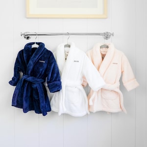 May include: Three plush robes hanging on a silver rack. The left robe is navy blue with the name henry embroidered on the chest. The middle robe is white with the name jacob embroidered. The right robe is light pink with the name alexa embroidered.