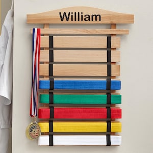 May include: A light-coloured wooden martial arts belt display rack with the name William engraved at the top. The rack features horizontal slats held by black fabric straps, displaying blue, green, red, yellow, and white belts, with a medal hanging.
