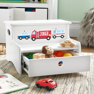 May include: A white wooden step stool with a storage drawer featuring a graphic of a blue police car, a red fire truck with the name Jonah printed on the side, and a white ambulance. The drawer contains a plush monkey and colorful toy balls.