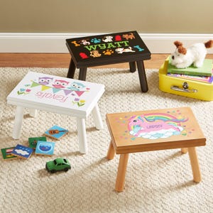 May include: Three small wooden step stools with four legs each. One black stool features the name WYATT with dog and fire hydrant graphics. One white stool features the name SYDNEY with owl and bunting graphics. One natural wood stool features the name LINDSEY with a unicorn graphic.
