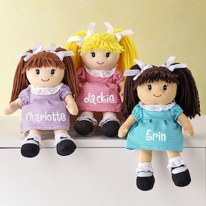 May include: Three plush dolls with yarn hair and white bows. The left doll has brown hair and a purple striped dress with the name Charlotte. The center doll has yellow hair and a pink striped dress with the name Jackie. The right doll has brown hair and a teal striped dress with the name Erin.