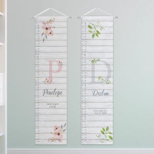 May include: Two vertical growth charts with a white wood plank background. The left chart features a pink letter P, floral accents, and the name Penelope with the text watch me grow 6.12.26. The right chart has a grey letter D, green leaves, and the name Declan with the text Sweet Baby Boy July 17, 2026. Both include imperial measurements.
