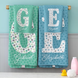 May include: Two personalized bath towels hanging on a rack. The left towel is teal with green and gray polka dots and the letter G. The right towel is blue with gray and blue stars and the letter E. Both feature the names Gabriel and Elizabeth.