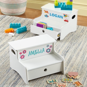 May include: Two white wooden step stools with storage drawers. One stool features the name LOGAN in blue with colourful vehicle decals. The other stool features the name AMELIA in teal with pink and blue flower decals. Both have side handle cutouts.