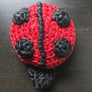 May include: A handmade, crocheted ladybug with a red body and black accents. The ladybug features black spots and a black stripe down its back. The antennae are also black. The craft item is made of yarn.