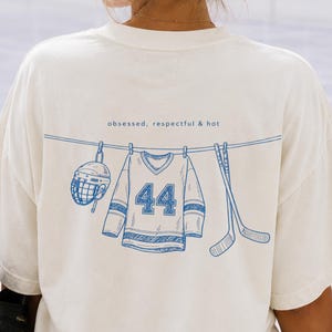 May include: An off-white short-sleeve t-shirt featuring a blue line drawing on the back. The graphic depicts a hockey helmet, a jersey with the number 44, and two hockey sticks hanging on a clothesline with the text obsessed, respectful & hot.