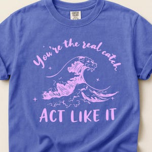 Könnte beinhalten: Ein blaues T-Shirt mit kurzen Ärmeln und einer hellrosa Grafik einer brechenden Meereswelle. Der Text "You're the real catch" ist in einem Bogen über der Welle gedruckt, und "ACT LIKE IT" steht in einer geraden Zeile unter der Welle.