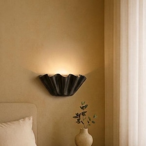Könnte beinhalten: Ein Schlafzimmer mit einer schwarzen, wellenförmigen Wandleuchte, die warmes Licht ausstrahlt. Das Bett ist mit beigefarbener Bettwäsche bezogen. Ein Nachttisch aus Holz zeigt eine Vase mit Grün und Büchern, was eine gemütliche Atmosphäre schafft.
