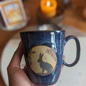 May include: A dark blue ceramic mug with a handle. The mug features a circular design with a silhouette of a rabbit in front of a full moon. The mug has a speckled finish and a rustic aesthetic.