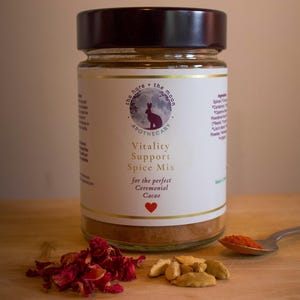 Op de afbeelding: Een glazen pot "Vitality Support Spice Mix" met een donkere dop, een etiket met een konijn- en maanlogo en de tekst "for the perfect Ceremonial Cacao". Een kleine hoop gedroogde rozenblaadjes, kardemompeulen en een lepel kruiden liggen voor de pot.
