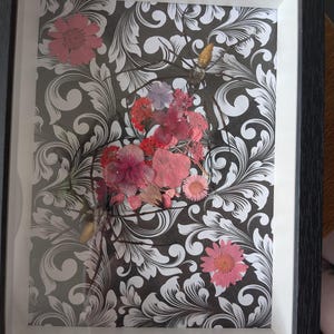 May include: A rectangular black frame containing a black and white damask patterned background. Pressed pink and red flowers are arranged in the centre, accompanied by two preserved spiders with long, thin legs and gold-toned bodies.