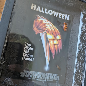 May include: A framed Halloween movie poster featuring a hand holding a knife against a carved pumpkin background. The text reads HALLOWEEN and The Night He Came Home. The black frame is decorated with black lace and artificial spider webs.
