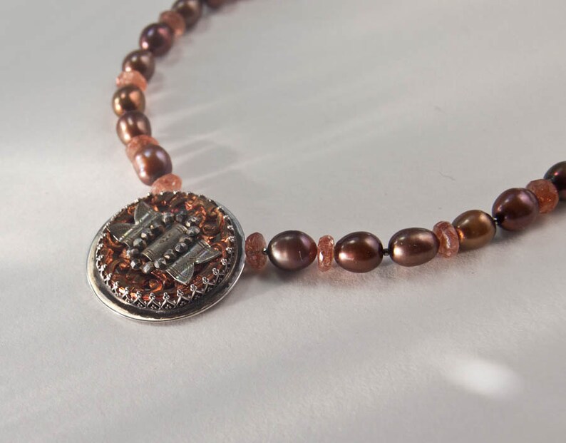 May include: A brown pearl and sunstone necklace with a silver and copper pendant featuring a geometric design.