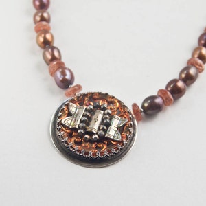 May include: A brown and silver necklace with a round pendant featuring a detailed design. The pendant has a copper-coloured centre with a silver design and a silver border. The necklace is made of brown and silver beads.