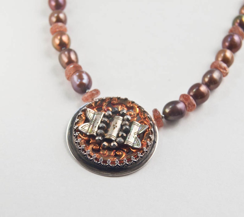 May include: A brown and silver necklace with a round pendant featuring a detailed design. The pendant has a copper-coloured centre with a silver design and a silver border. The necklace is made of brown and silver beads.