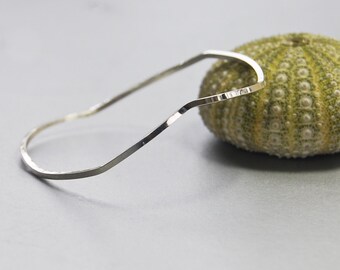 May include: A silver bangle bracelet with a wavy, textured design. The bracelet is open and has a simple, minimalist style.