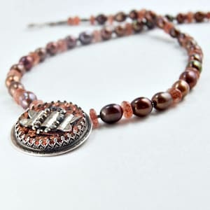 May include: A brown and silver necklace with a round pendant featuring a copper and silver design. The necklace is made with brown pearls and small pink stones.