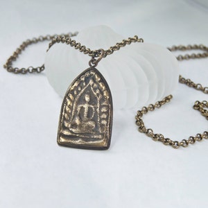 May include: A gold-toned necklace with a pendant featuring a detailed image of a seated Buddha figure. The pendant is shaped like a pointed arch and has intricate designs around the Buddha.