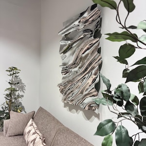 Puede incluir: Una escultura de pared rectangular de metal con una superficie plateada pulida altamente reflectante, con ondas profundas y contornos orgánicos fluidos que crean reflejos abstractos y distorsionados del entorno circundante.