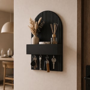 May include: A black wall-mounted organiser with an arched, ribbed back panel and a rectangular shelf. The shelf holds a beige vase with dried grasses, a small marble dish, and a glass reed diffuser. Four metal hooks hang keys below the shelf.